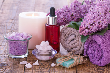 Fototapeta na wymiar Bowls with sea salt, burning candle, red bottle with aromatic oil, handmade soap, lilac flowers and towels for bathroom procedures