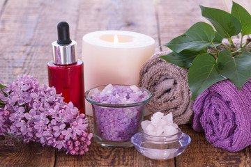 Obraz na płótnie Canvas Lilac flowers, red bottle with aromatic oil, burning candle, bowls with sea salt and towels with green leaves on wooden boards