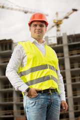 Smiling young man in red hardhat and green safety vest standing on building site