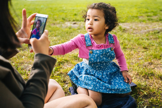 Unrecognizable mixed race mother taking a photo her a three years old dark skinned cute baby girl with smartphone in grass at summer park. Digital family memories concept.