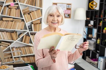 Difficult language. Cheerful mature woman carrying glasses and reading book