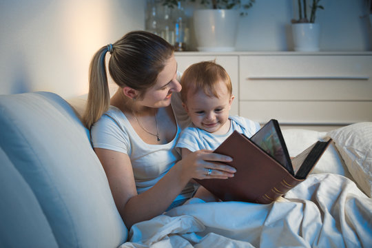 Happy smiling baby boy sitting in bed with mother and looking on pictures at big book