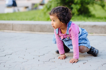 Fototapeta premium Dark skinned hispanic toddler girl crawling outdoor. Caucasian child dressed in casual clothing: blue denim sarafan, pink shirt and sneakers. portrait of kid with beautiful dark curly hair.