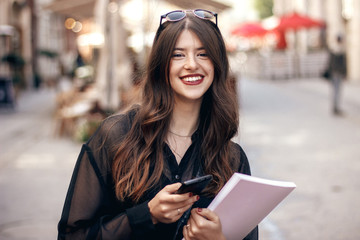 beautiful stylish hipster girl texting on phone, smiling in sunny city street, holding magazine. happy gorgeous woman, dressed in fashionable outfit, using smartphone and enjoying time