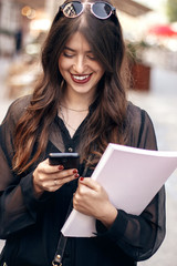beautiful stylish hipster girl holding phone and magazine, walking and smiling in sunny street. happy gorgeous woman, dressed in fashionable outfit, enjoying time in european city