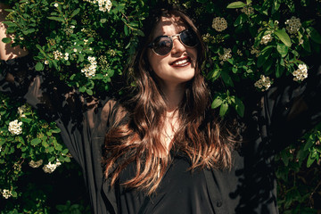beautiful Stylish Hipster Girl with beautiful hair relaxing at floral bushes in sunny day. Gorgeous Boho Woman with sunglasses in fashionable outfit enjoying day in flowers in garden