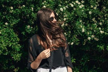 beautiful Stylish Hipster Girl with beautiful windy hair having fun at floral bushes in sunny day. Boho Woman with sunglasses in fashionable outfit, smiling and enjoying day in garden