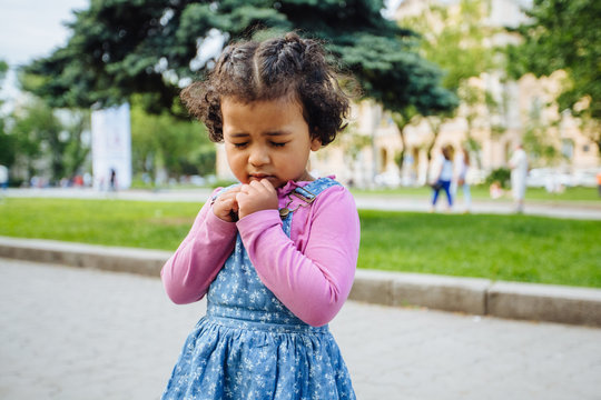 Dark Skinned Baby Toddler Girl Praying With Hands Folded And Eyes Closed Outside In City Square Or Park Background.