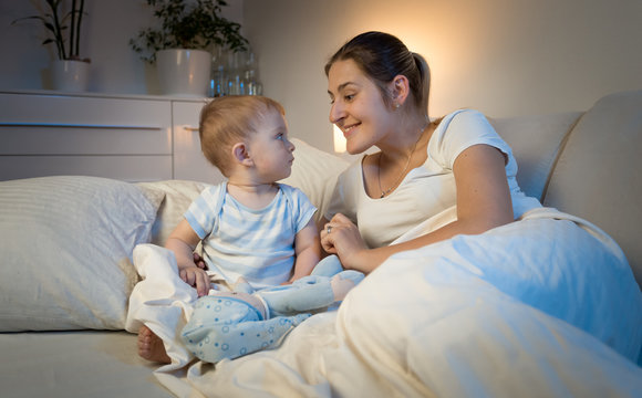 Portrait of smiling young mother and baby boy lying in bed at night and looking at each other