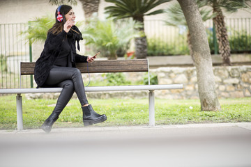 ragazza seduta in una panchina sorride con cuffie con bandiera UK ascolta la musica al parco  e fa il gesto di OK 