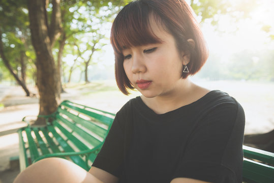 Young Asian woman sitting alone on the public bench in the park surrounded with nature and warm sunlight. Young lonely woman sitting alone in the park. Outdoor activity in the park concept.