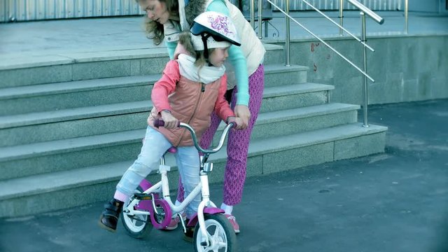 A laughing, smiling mother pushes her daughter forward on a warm spring day, when she teaches her to ride a bike along the city's sidewalk near a green park.