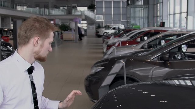 Young male employee is closing hood of car, standing in auto salon, bearded guy in work process in modern automobile center with rows of new cars. slow motion. Seller is dressed in white shirt with
