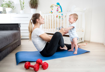 Cute 9 months old baby boy exercising with young mother on fitness mat at home