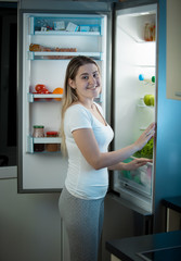 Portrait of smiling young woman in pajamas standing at open refrigerator at home