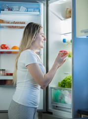 Smiling hungry woman taking sweet donut out of refrigerator at night