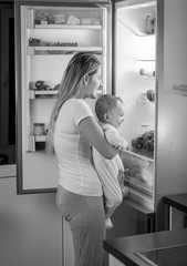 Fototapeta na wymiar Black and white image of mother with cute baby looking for something to eat in refrigerator at night