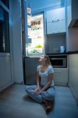 Fototapeta na wymiar Young hungry woman sitting on kitchen floor at night and looking on the fridge