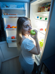 Portrait of hungry young woman eating fresh green apple on kitchen at night