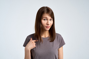 a Studio portrait of an attractive brunette woman in a gray t-shirt with uncertainty on her face showing her finger at herself. 