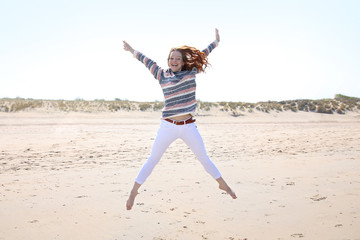 Hübsche rothaarige Frau an einem Strand springt vor Freude mit ausgebreiteten Armen in die Luft