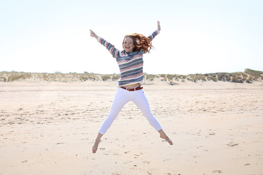 H&uuml;bsche rothaarige Frau an einem Strand springt vor Freude mit ausgebreiteten Armen in die Luft