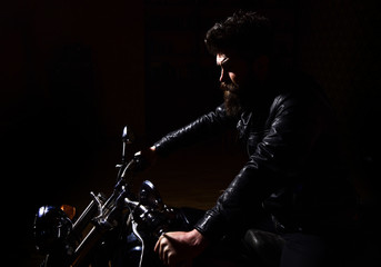 Masculinity concept. Man with beard, biker in leather jacket sitting on motor bike in darkness, black background. Macho, brutal biker in leather jacket riding motorcycle at night time, copy space.
