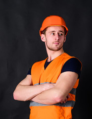 Man in helmet, hard hat hold arms crossed on chest, black background. Worker, contractor, builder on strict face with muscular biceps. Builder in helmet posing confidently. Strong builder concept.
