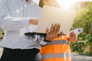 Asian man and woman engineer with the blue safety helmet meeting at the construction site with blueprints and laptop.