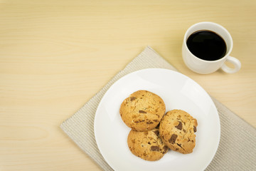 a cup of coffee, three pieces of chocolate chip cookies in a white round dish on the wooden table.