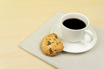 a cup of coffee and chocolate chip cookies in a white round dish on the wooden table.