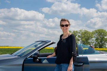 Attractive smiling young woman standing next  luxury convertible car. she looks at camera. 