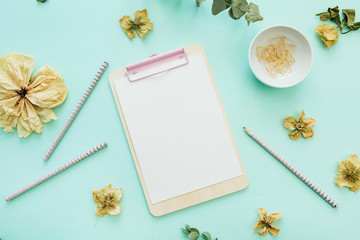Flat lay office workspace with blank clipboard, flowers and branches on blue background. Top view minimal mock up template concept.