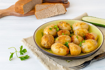 Young fried potatoes in a round  plate, next to a fresh cucumber and bread on an old wooden table. 