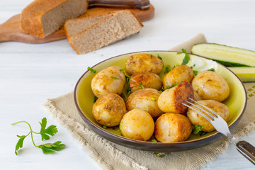 Young fried potatoes in a round  plate, next to a fresh cucumber and bread on an old wooden table. 