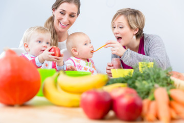Obraz na płótnie Canvas Two happy mothers and best friends smiling while feeding together their healthy baby girls at table with natural and nutritive food indoors at home