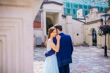 Beautiful newlyweds posing in old town. Adorable bride and groom kiss each other behind old orange wall under green tree leaves. Wedding day. Woman in blue dress. Man in suit. Young wedding couple.