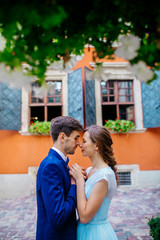 Beautiful newlyweds posing in old town. Adorable bride and groom kiss each other behind old orange wall under green tree leaves. Wedding day. Woman in blue dress. Man in suit. Young wedding couple.