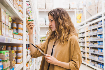 Young beautiful woman with a tablet selects baby food in a supermarket, the girl carefully reads the composition of the product