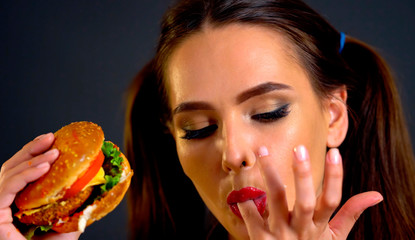 Woman eating hamburger. Girl wants to eat burger. How quickly to cook dinner. Portrait of person with good appetite have greedily dinner delicious sandwich on a black background. She licks her finger.