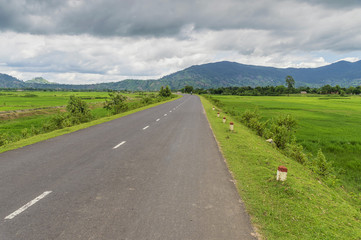 Fototapeta na wymiar road between rice fields against the backdrop of mountains covered with greenery, tropical vegetation, clouds, sky Daklak province, Vietnam