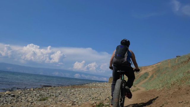 Fat bike also called fatbike or fat-tire bike in summer driving on the road. The guy rides by the hill on a sand clay path, behind him the shore by the sea. Slow motion shooting 180fps.