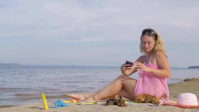 Woman with cell phone on the beach. She rewrites by phone, looks at photos, makes selfies. The girl is taking pictures of the sea on the phone. Two of her little dogs are resting nearby.