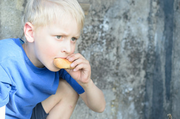 A hungry dirty boy greedily eats a crust of bread against the wall.