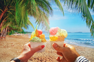 Couple with beautiful bright  sweet Italian ice-cream with different flavors  in the hands  . Background of  panoramic view of tropical beach with coconut palm trees. Koh Samui, Thailand