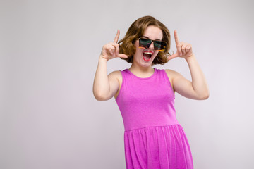 Charming young girl in pink dress on gray background. Cheerful girl in square glasses. The girl points fingers at the top