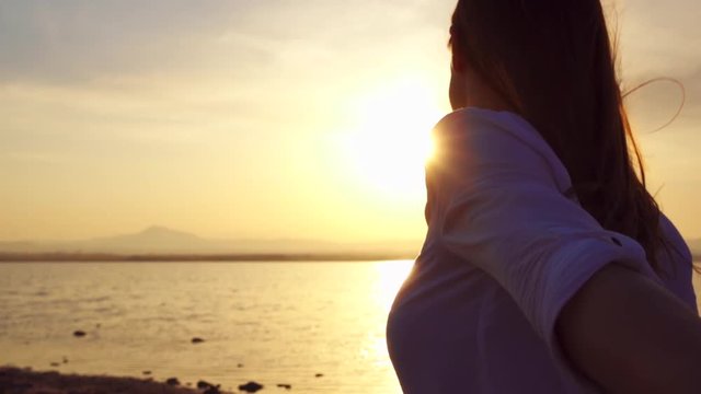 Young couple holding hands outdoors at golden hour at sunset. Happy woman in white shirt leading her boyfriend on lake in slow motion. Female traveler turning around and smiling