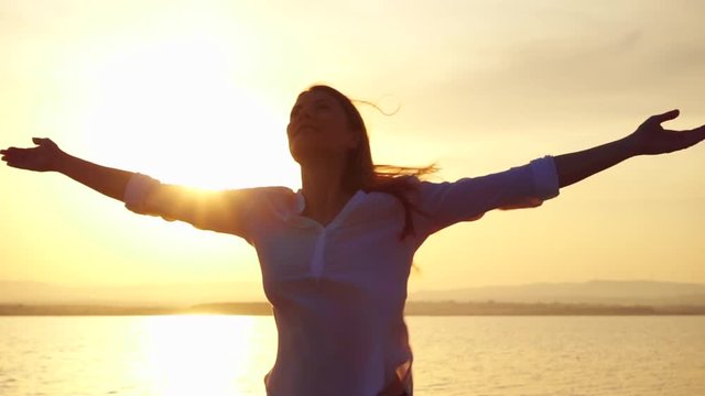 Dark silhouette of young woman raising arms up at sunset on lake. Female figure outstretching hands at golden hour in slow motion