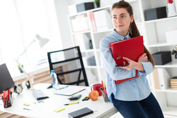A young girl in the office is standing, leaning on a table, and is holding a phone and a folder with documents.