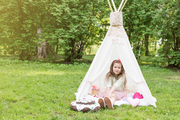 Little girl lying and playing in a tent, children's house wigwam in park. Happy mothers day. Child with big donuts and straw hat and heart-shaped pillows. Summer, happy childhood concept. Cute © MartaKlos